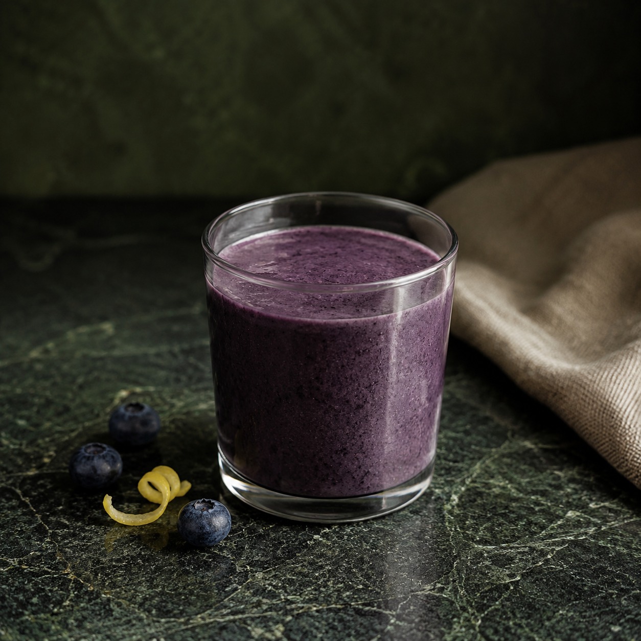 A small deep-purple blueberry lemon protein smoothie in a clear glass with blueberries and a curl of lemon peel.
