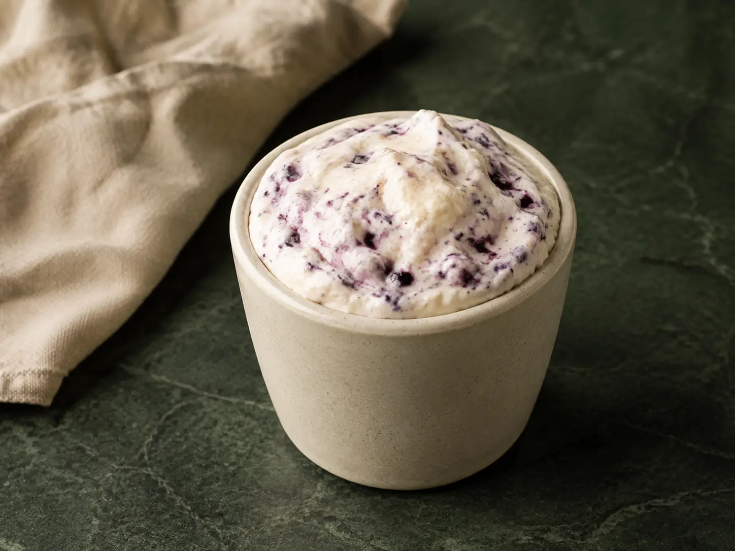 Pale lemon cheesecake fluff with crushed blueberries in a small cream stoneware cup.
