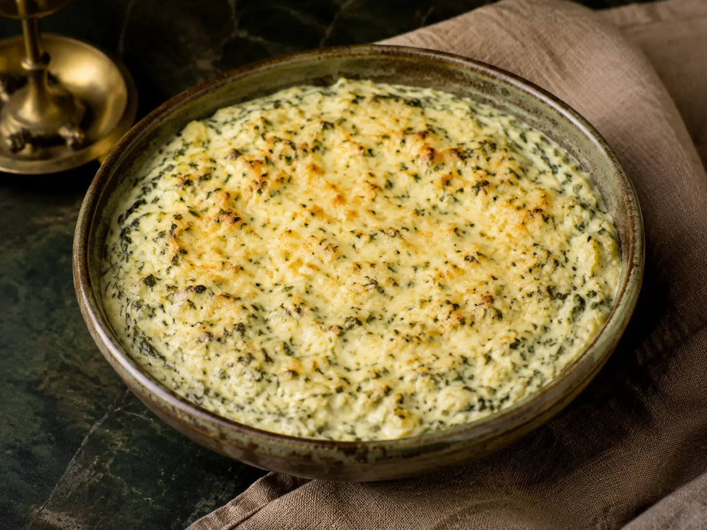 A small rustic bowl of warm creamy spinach and artichoke dip with fine green flecks and a lightly golden surface.