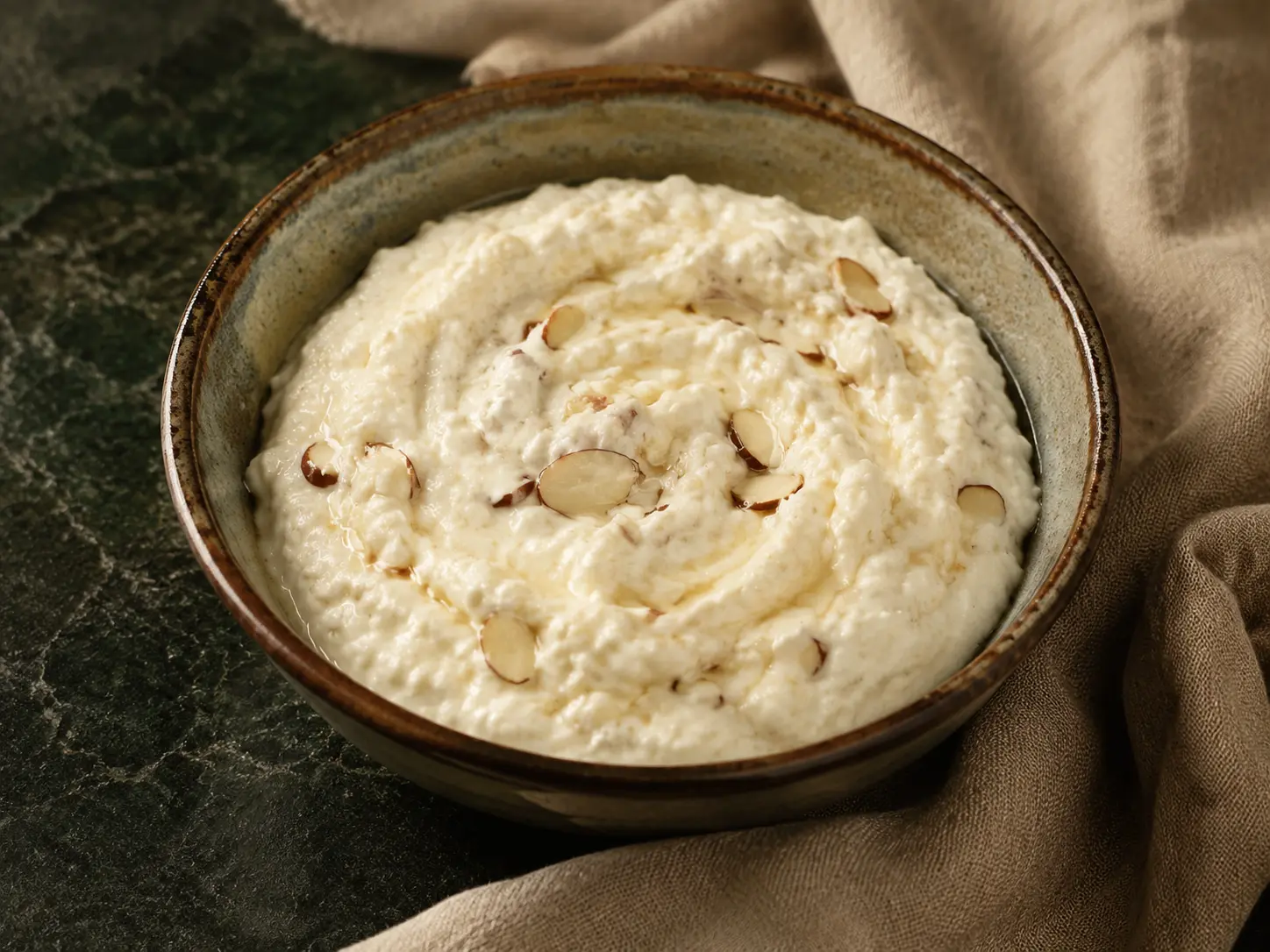 A rustic bowl of creamy honey cottage-cheese dip with sliced almonds on dark forest-green stone beside warm linen.
