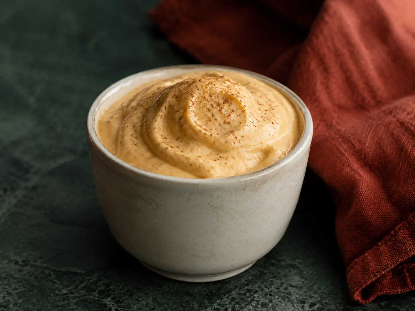 Pumpkin cheesecake protein fluff dusted with cinnamon in a small cream stoneware cup.