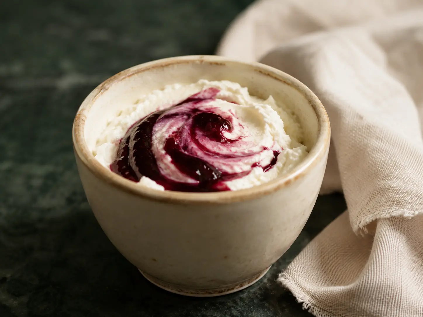 A small cream cup of whipped ricotta with a smooth seedless berry swirl on dark forest-green stone.