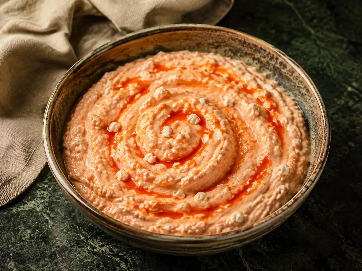 A rustic bowl of coral-pink sriracha-feta cottage-cheese dip with fine white feta flecks on dark green stone.